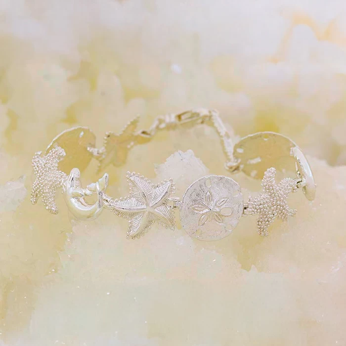14K white gold bracelet featuring starfish, dolphins, and sand dollar charms on a light sandy background.